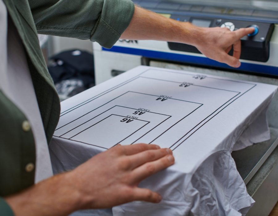 Cropped view of young artisan using blurred screen printing machine near t-shirt with marking while working in print studio, customer-focused small business concept